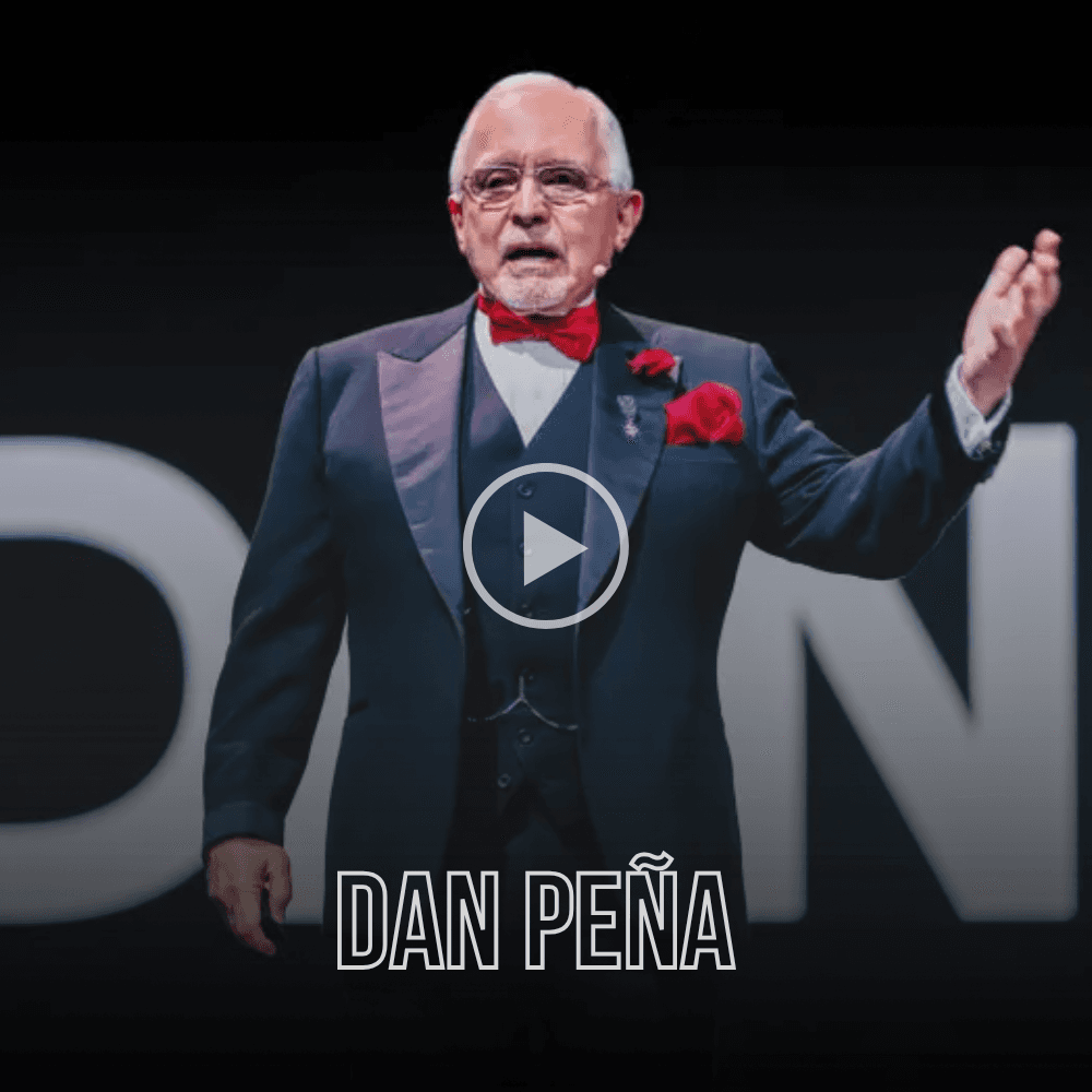 Dan Peña wearing a tuxedo and red bow tie, gesturing during a stage presentation.