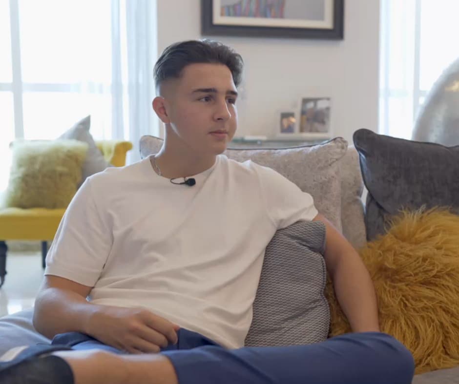 Young man with a lapel microphone sits on a couch with grey and yellow pillows.