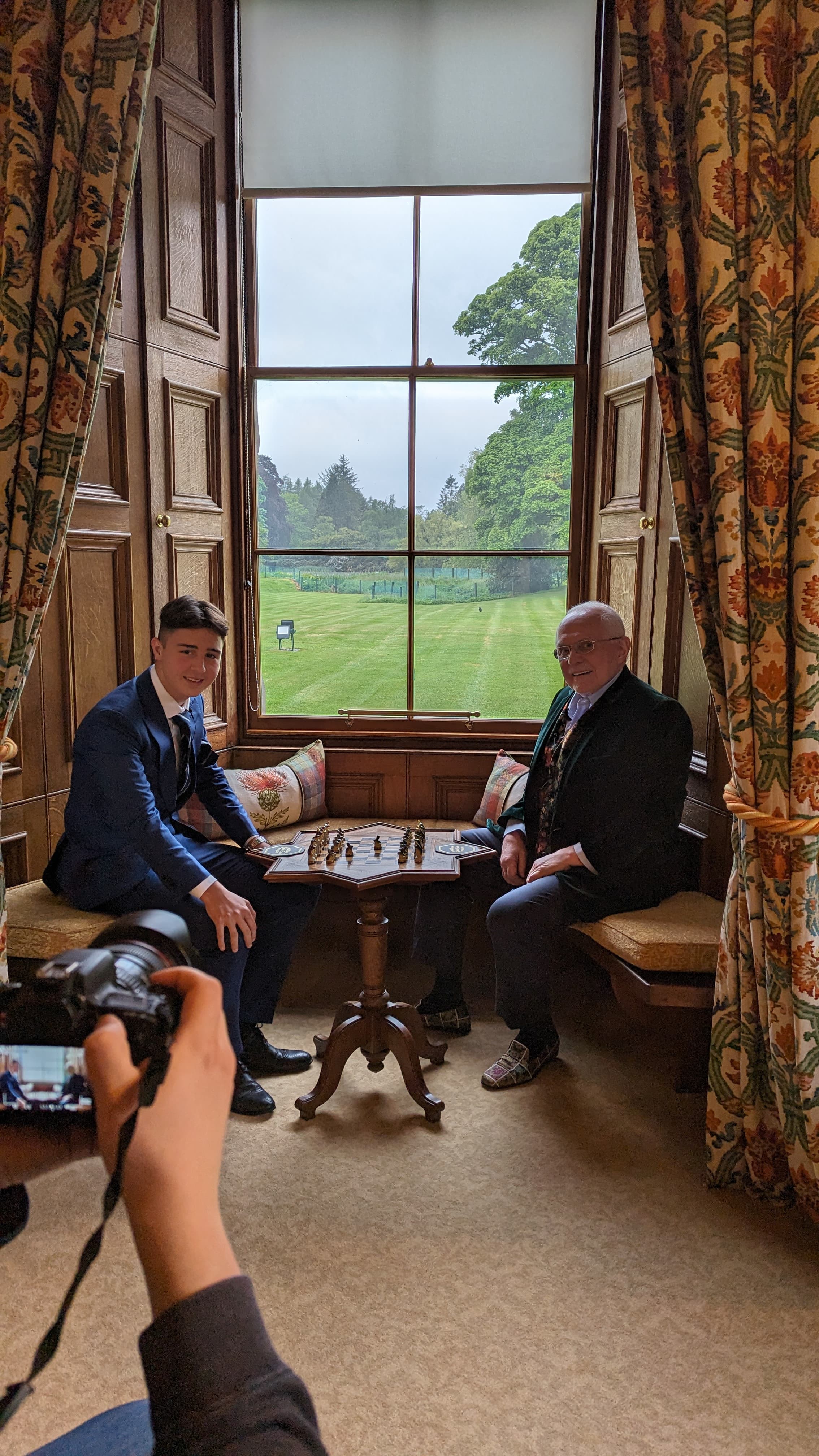 Two men pose at a chessboard in a window nook overlooking a green lawn.