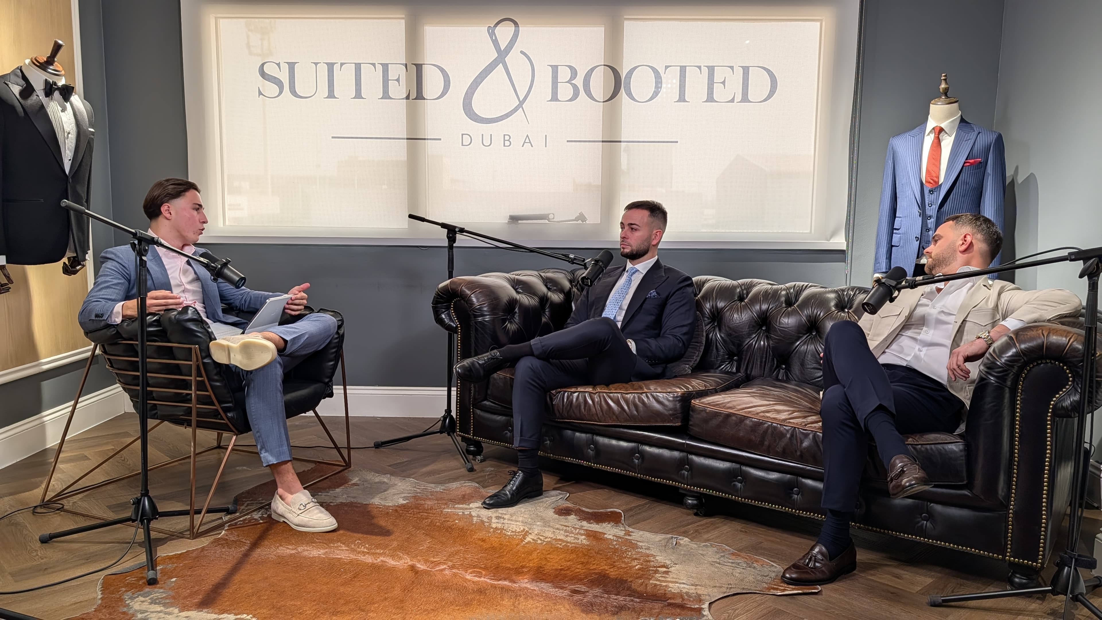 Three men in suits record a podcast in a studio featuring Suited & Booted signage.