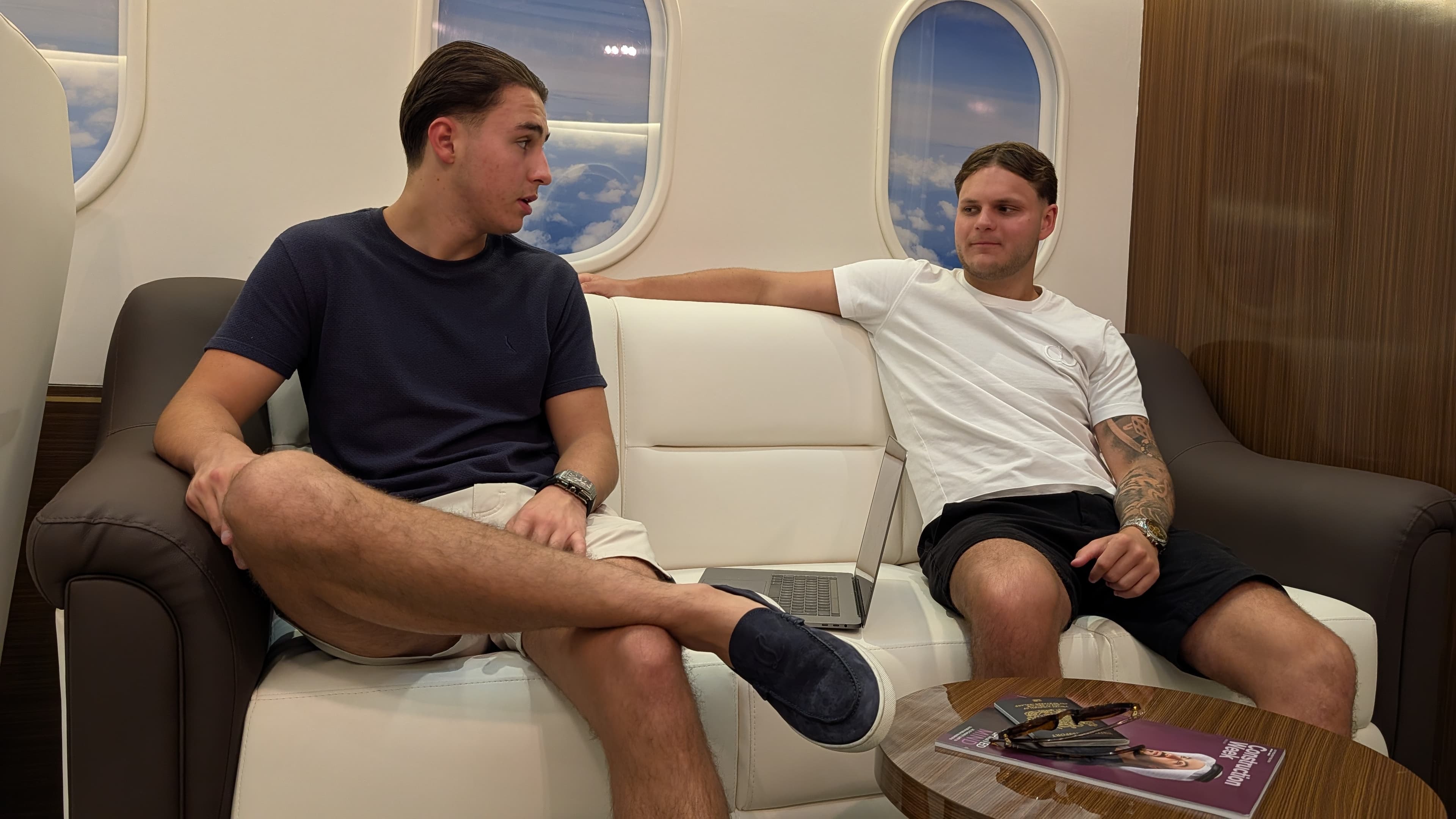 Two young men converse on a white leather sofa inside a luxurious private jet cabin.