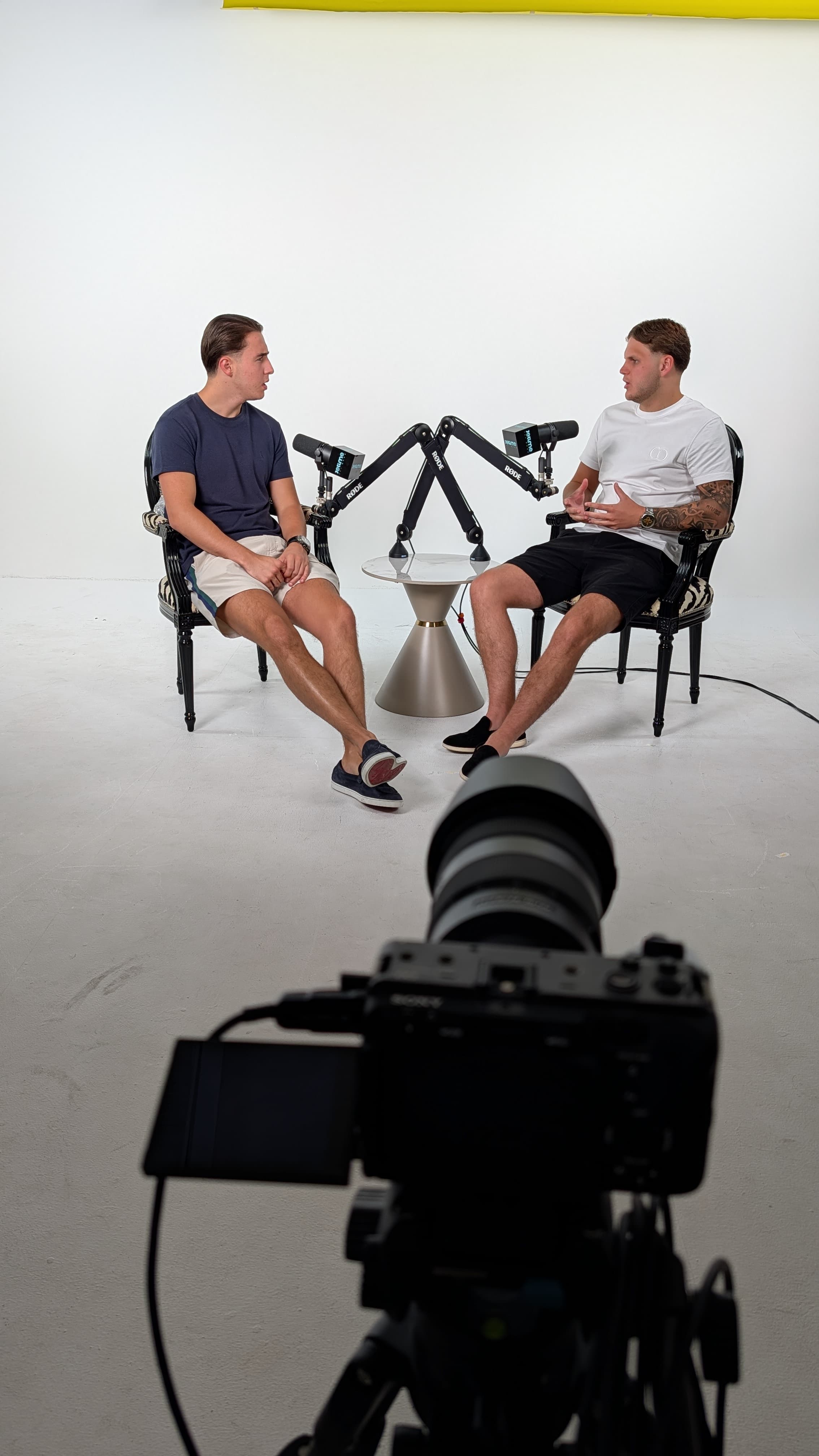 Two men recording a podcast in a white studio, viewed from behind a video camera.