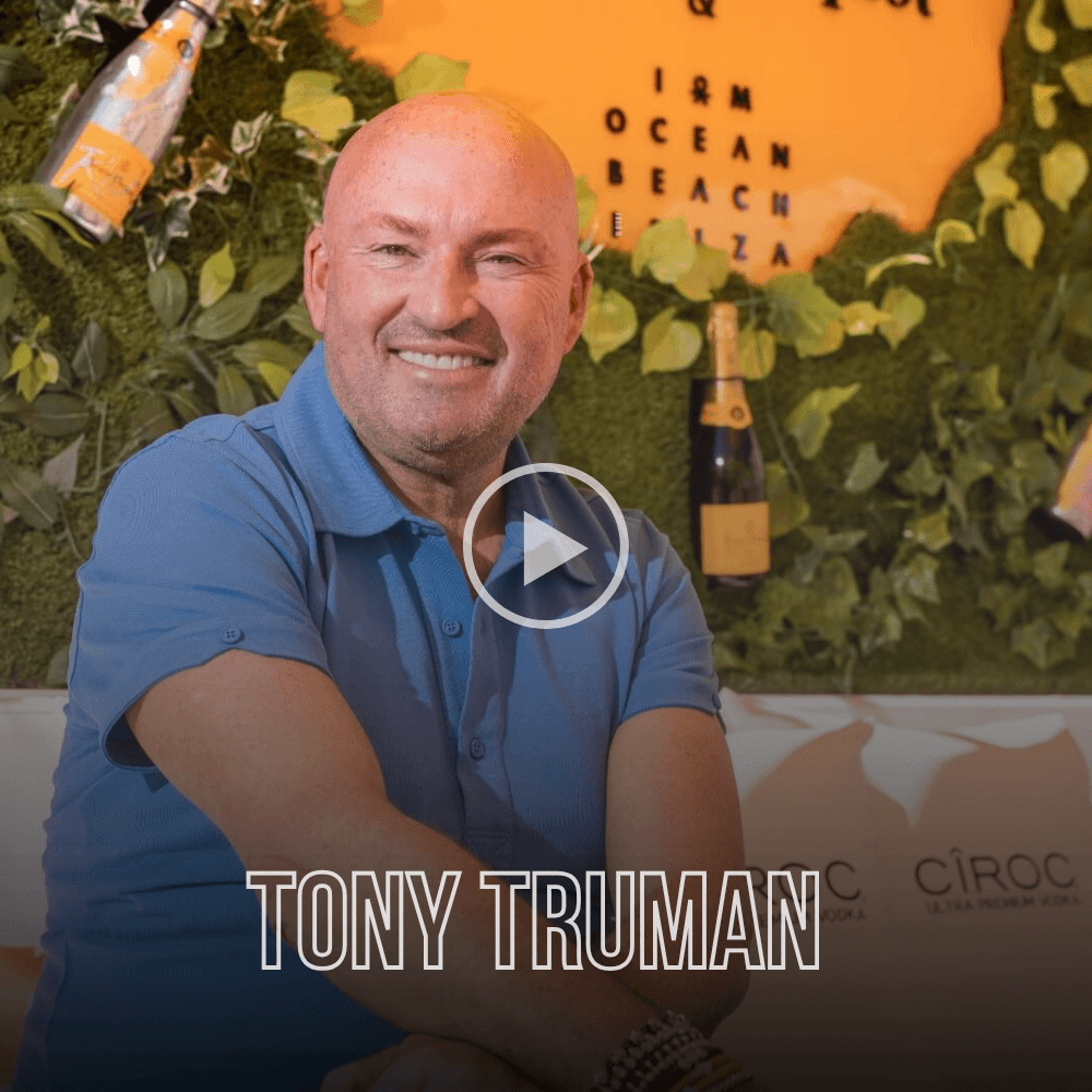 Tony Truman smiling in a blue polo shirt against a green wall with champagne bottles.